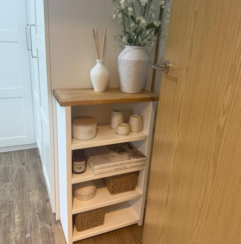 White bookshelf with wooden top, decorated with a vase of flowers, reed diffuser, jars, and woven baskets.