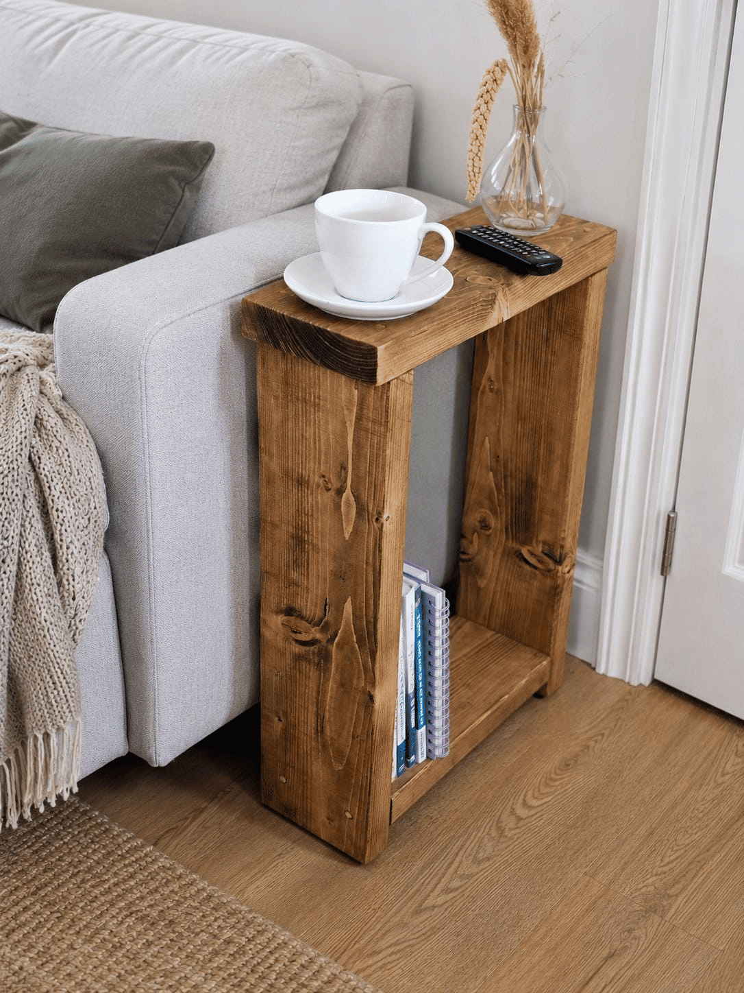 Slim dark wooden side table beside a grey sofa, with a cup on top and books on the lower shelf.