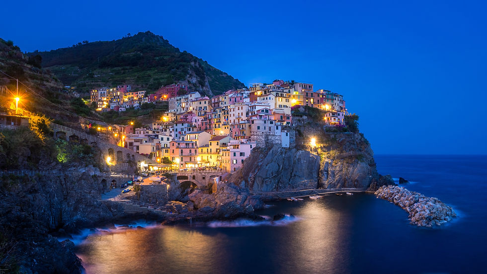 Colorful hillside village at dusk by the sea, with glowing lights reflecting on the water, set against a deep blue sky and rocky cliffs.