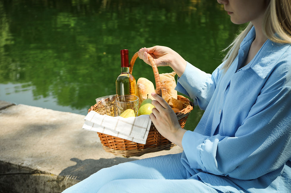 Woman in blue sits by a lake with a picnic basket containing bread, wine, glasses, and fruit. Calm, sunny setting with greenery reflected in water.