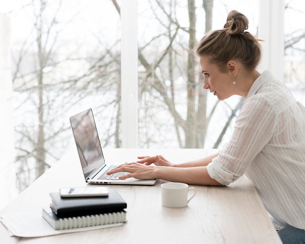 Woman in a white shirt works intently on a laptop at a wooden table. Background shows bare trees through large windows, creating a calm mood.