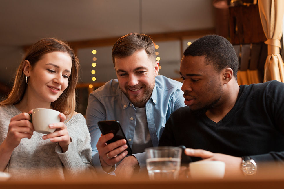 Three people smile and look at a phone in a cozy cafe setting with warm lighting. One holds a white cup. Relaxed and cheerful mood.