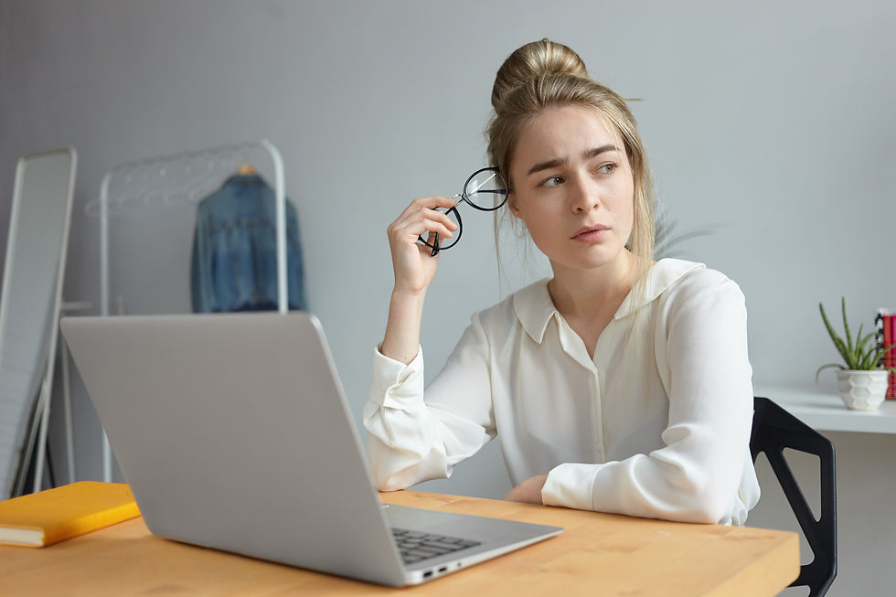 Woman in a white shirt sits at a desk with a laptop, holding glasses, looking pensive. Background includes a jacket, books, and a plant.