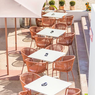 Outdoor cafe with white tables, brown wicker chairs, and beige umbrellas. Bright, sunny setting with potted plants and a colorful sculpture.