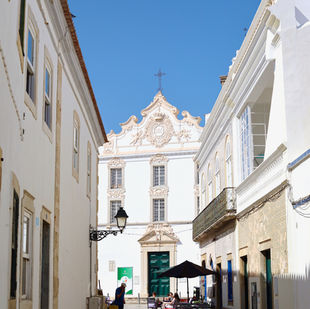 Sunny street scene with people seated at outdoor café tables, surrounded by white historic buildings and ornate church facade under a blue sky.