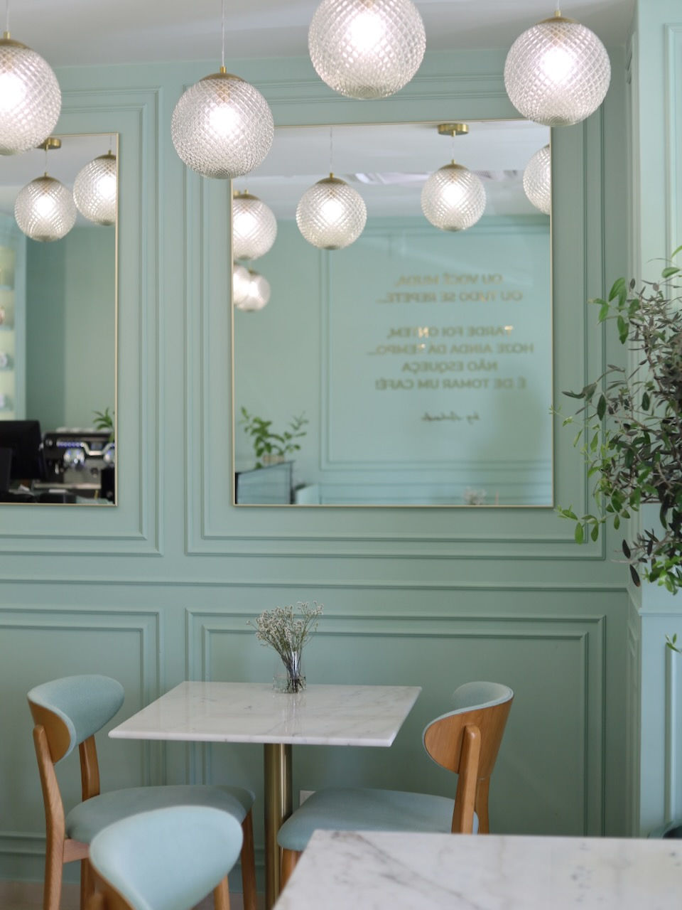 Elegant café interior with mint-green walls, a marble table, and wooden chairs. Glass pendant lights hang above, and a mirror reflects text.