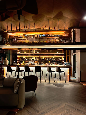A bartender works behind a well-lit yellow golden bar with high stools, bottles, and glasses. Modern decor with warm lighting creates a cozy ambiance.