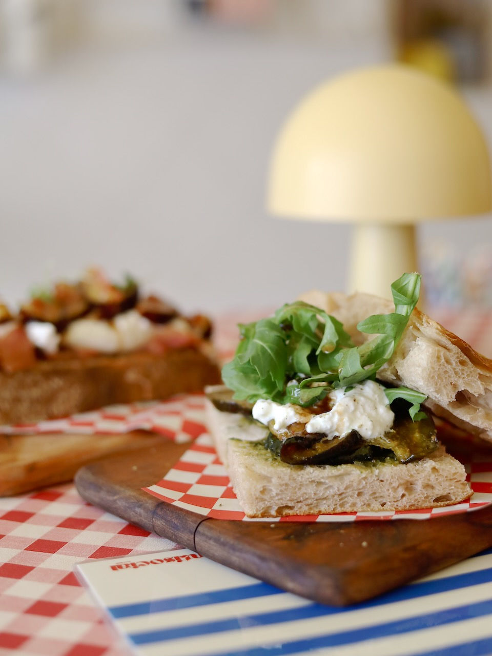 Open sandwich with arugula and cream cheese on focaccia sits on a wooden board with red checkered paper. Soft, warm lighting from a nearby lamp.