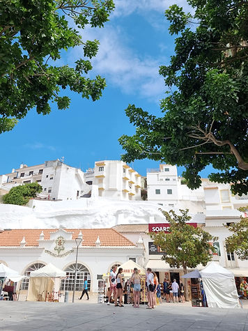 Narrow cobblestone street with white buildings; distant orange manor on green hill. Lamppost in foreground. Sunny, serene atmosphere.