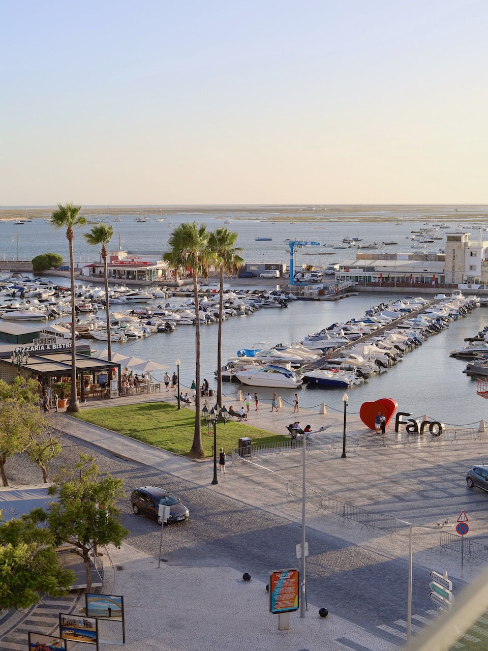 A marina with many boats in Faro at sunset, palm trees, people walking, a "Faro" sign with a red heart, and a pizzeria nearby.