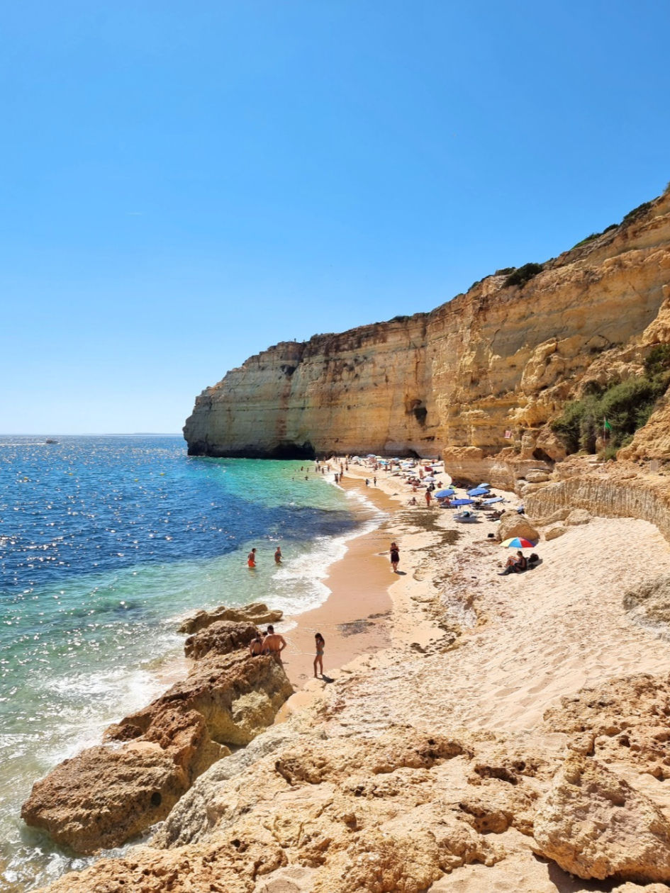 Sunlit beach with cliffs, swimmers, and people relaxing on sand. Clear blue sky and sea; colorful umbrellas create a vibrant, lively setting.