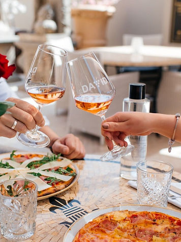 A couple cheering with 2 rosé glasses over their 2 pizzas. The ambiance is casual and elegant
