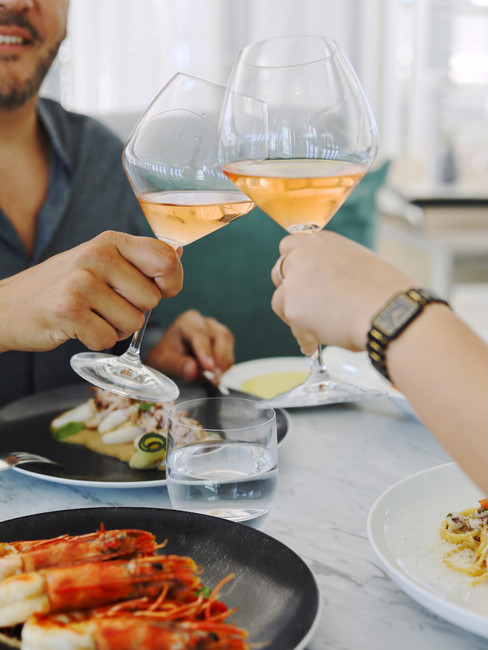 Two glasses of Rose wine cheering, below the glasses are dishes with Italian food at a restaurant in Algarve