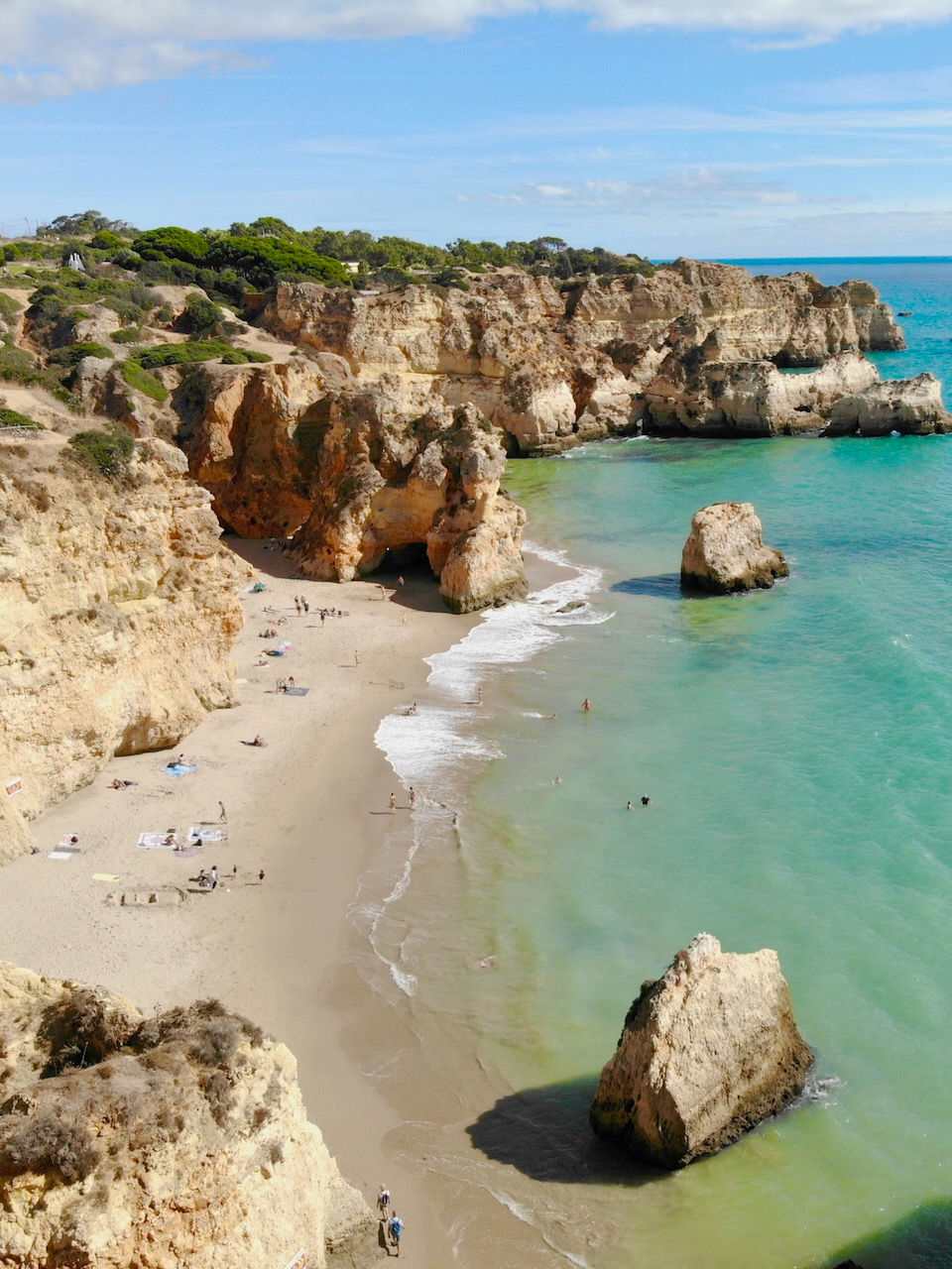 Cliff-lined beach with people sunbathing and swimming in turquoise waters. Bright sky, rocky cliffs, and beach towels create a peaceful scene.