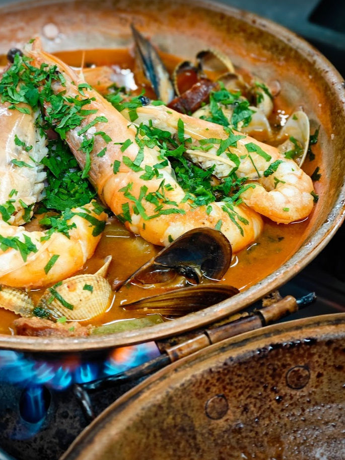Seafood stew in a copper pan, featuring prawns and mussels. Garnished with fresh herbs on a blue flame. Warm, hearty ambiance.
