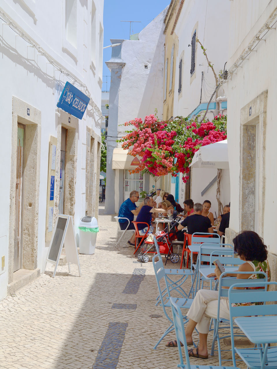Narrow cobblestone alley in bright sunshine, with people dining at tables. Vibrant bougainvillea blooms overhead, creating a lively, cozy atmosphere.
