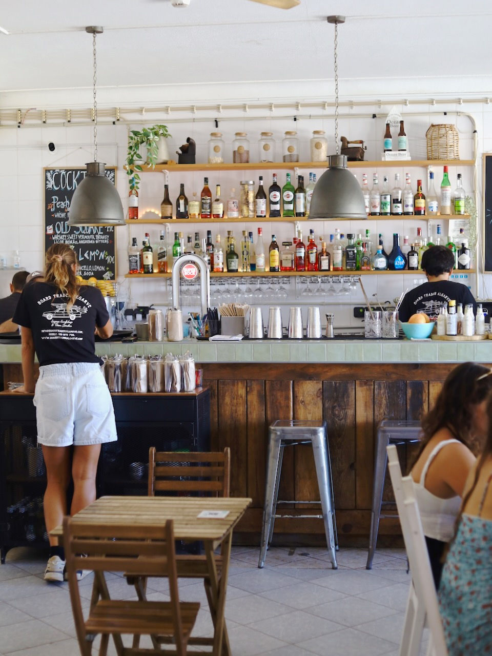 Cafe bar with wooden decor, bottles on shelves, two staff in black shirts working. Menu text on chalkboard. Casual and lively mood.