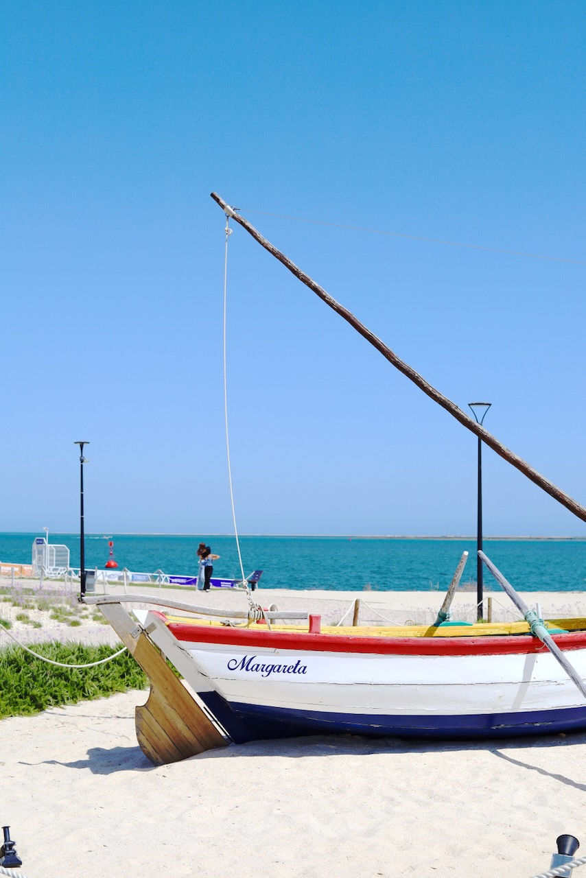 A colorful boat named rests on a sandy beach in the Algarve with a clear blue sky and ocean. A person stands near the water, embracing.