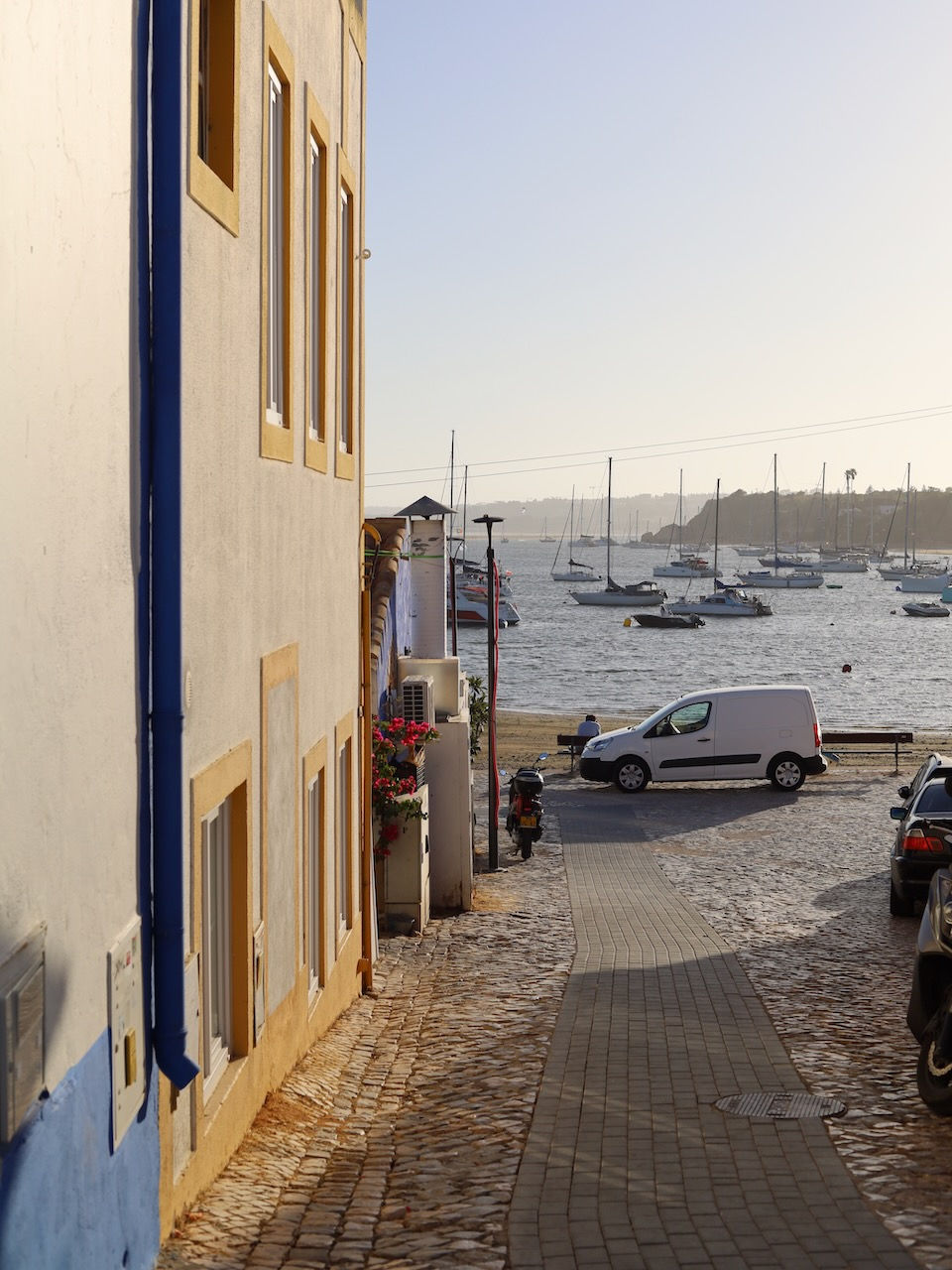 Cobblestone street in Alvor leading to a harbor with boats. Colorful buildings line the street, and a white van is parked beside the water. Calm mood.