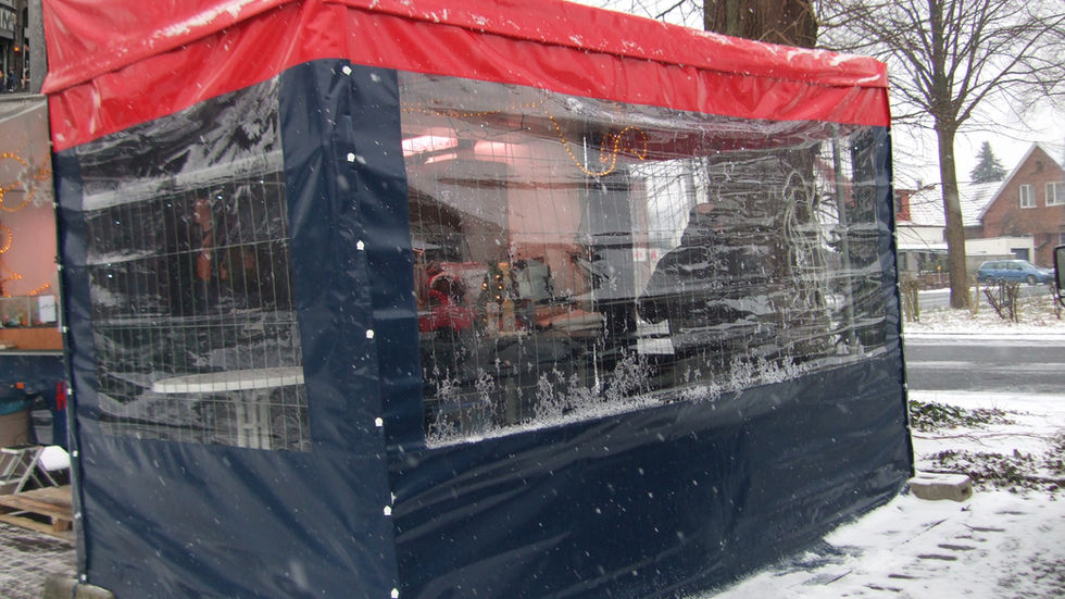 Ein rechteckiges Zelt mit rotem Dach und dunkelblauen Seitenwänden, die teilweise aus transparentem Kunststoff bestehen. Das Zelt steht auf schneebedecktem Boden, im Hintergrund sind Bäume ohne Blätter zu sehen.