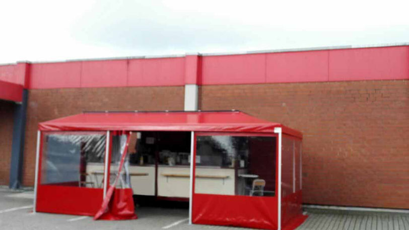 Rotes Imbiss-Anbauzelt mit transparenten Fenstern, offenem Vorhang am Eingang, davor gepflasterter Boden, im Hintergrund rotes Gebäude mit Backsteinwand.