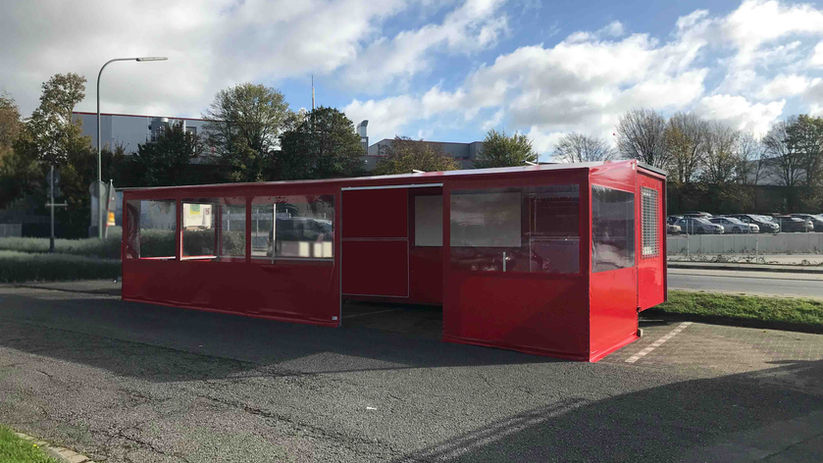 Rotes, rechteckiges Imbiszelt mit großen Fenstern auf Asphaltfläche, umgeben von Bäumen und geparkten Autos.