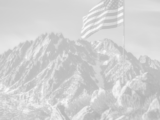 U.S. flag waves near rugged mountains in monochrome. Snow patches visible, rocky terrain in foreground. Calm and majestic setting.