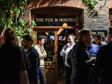People socialize outside The Fox & Hounds pub under ivy-covered brick, holding drinks in warm evening light.