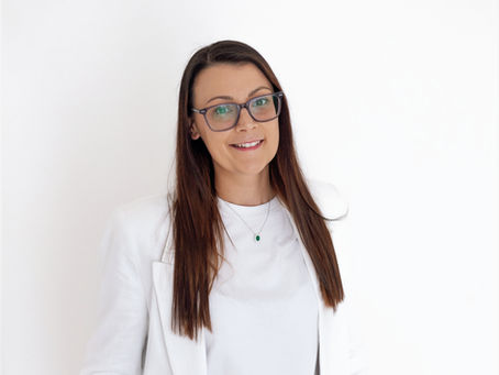 Woman with long hair and glasses smiling, wearing a white blazer and necklace, standing against a plain white background.