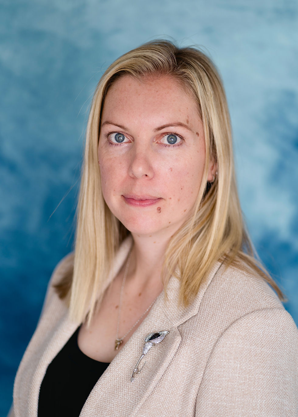 Blonde woman in beige blazer with brooch, neutral expression, blue textured background.