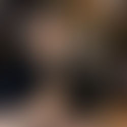 A smiling person in a dark shirt sits at a wooden table in a cozy, warmly lit cafe. Decorative lights and signs are visible in the background.