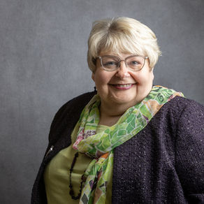 Smiling woman with glasses in a green scarf and dark jacket against a grey backdrop. She exudes a friendly and welcoming vibe.