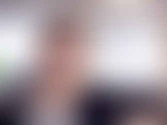 Smiling woman in checked shirt in bright office setting, blurred background with large windows, creating a welcoming atmosphere.