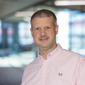 Smiling man in a light pink shirt stands indoors. Blurry office background with bright lighting.