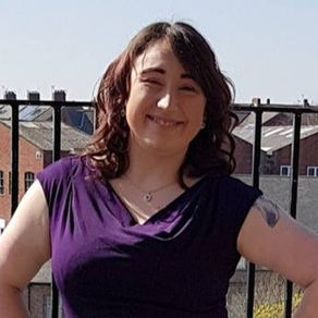 Smiling person in a purple shirt stands confidently outdoors. Background shows rooftops and a clear sky, conveying a cheerful mood.