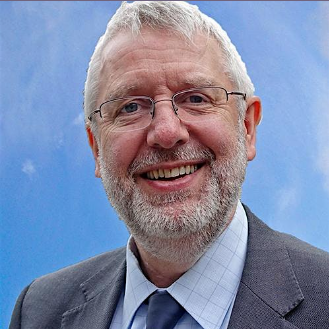 Smiling man with glasses and a beard in a grey suit and blue tie, against a clear blue sky background.