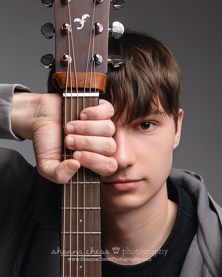 Close-up portrait of a young man holding a guitar neck over one eye, against a gray background, during his senior portrait photoshoot.
