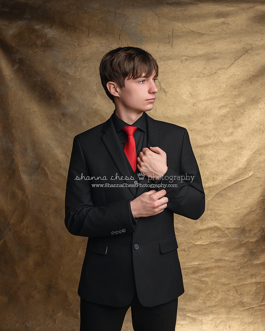 Teen boy in black suit and red tie adjusts his jacket cuff while standing against a textured tan studio backdrop in Eugene Oregon.
