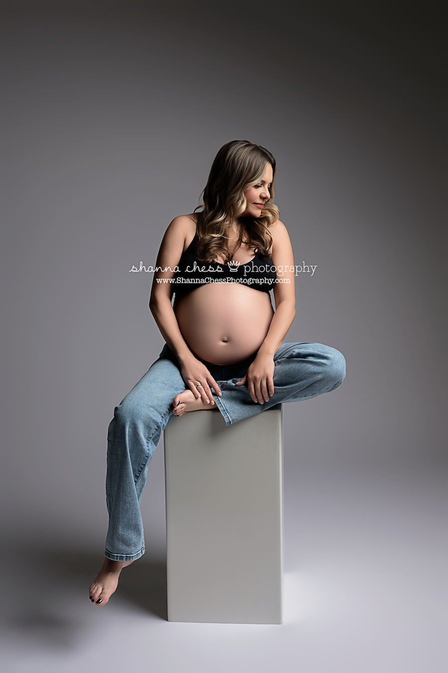 Pregnant woman in jeans and a black bra seated on a photographer's posing block with one leg tucked under her, gazing off to one side