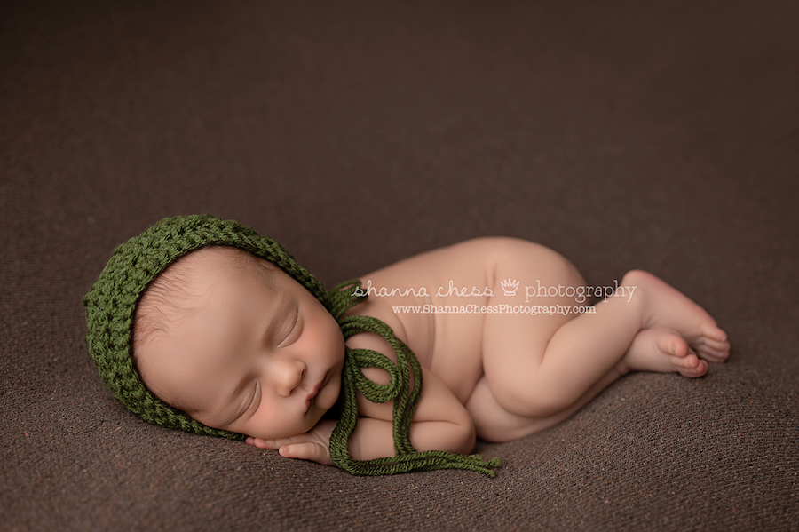 Newborn boy in soft crocheted green bonnet asleep on a twill brown beanbag.