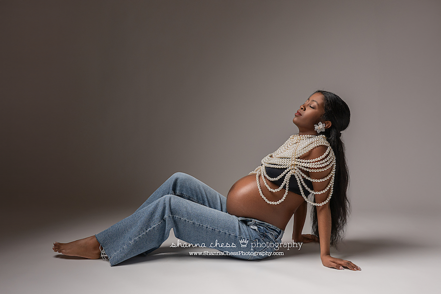 Pregnant woman in jeans and bead necklace bra top leans back on the floor of the studio with her eyes closed during her maternity photoshoot
