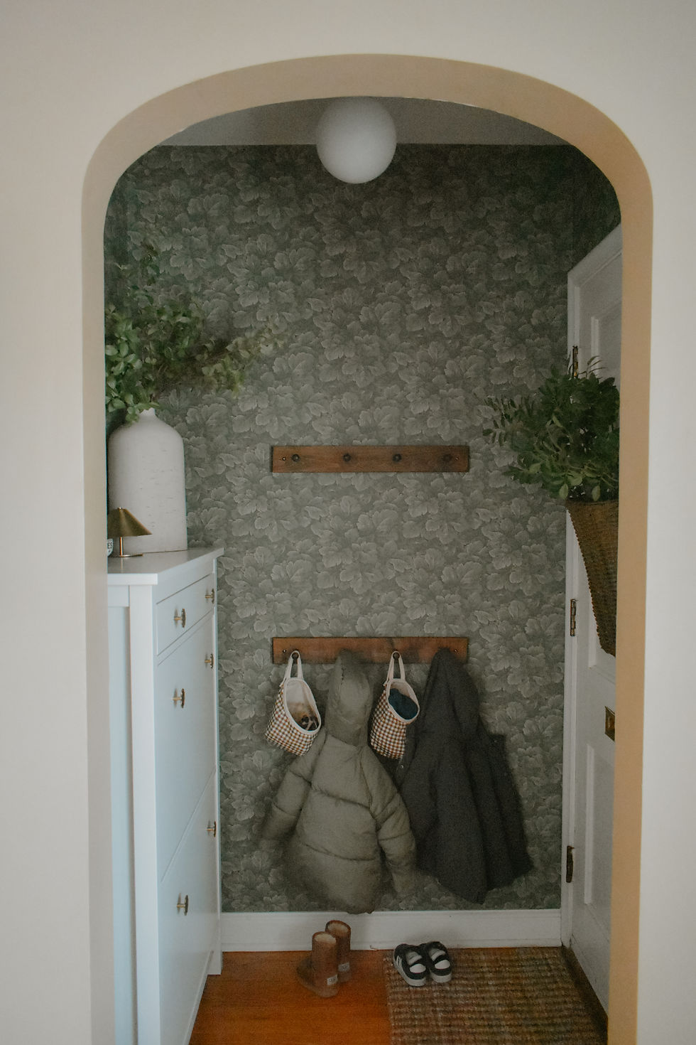 Cozy entryway with green floral wallpaper, coats on hooks, and checkered bags. A white cabinet with decor sits on the left. Relaxed ambiance.