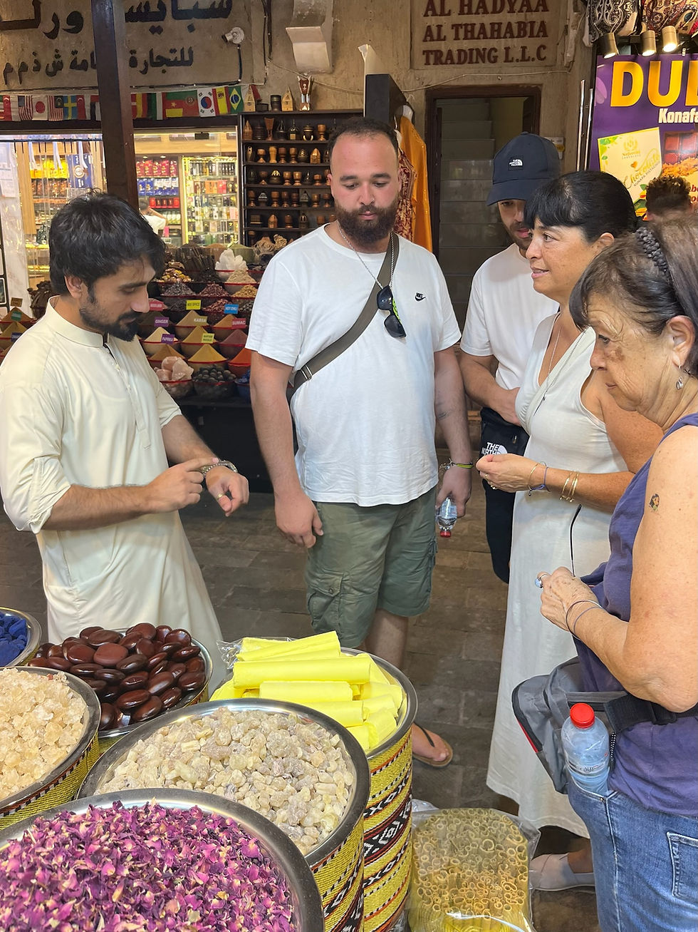 Tourists gather around a spice vendor in Dubai Spice Souk market, watching him demonstrate a bracelet amid colorful spice bowls.