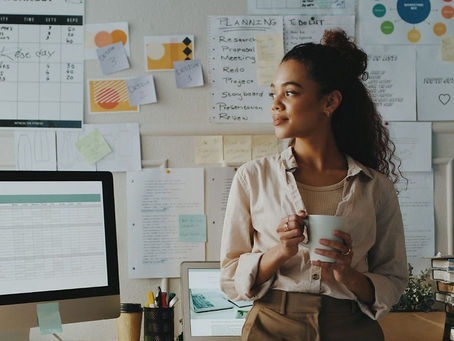 Empreendedora segura uma xícara enquanto sorri levemente em ambiente de trabalho home office com vários projetos fixados em papeis nas paredes.