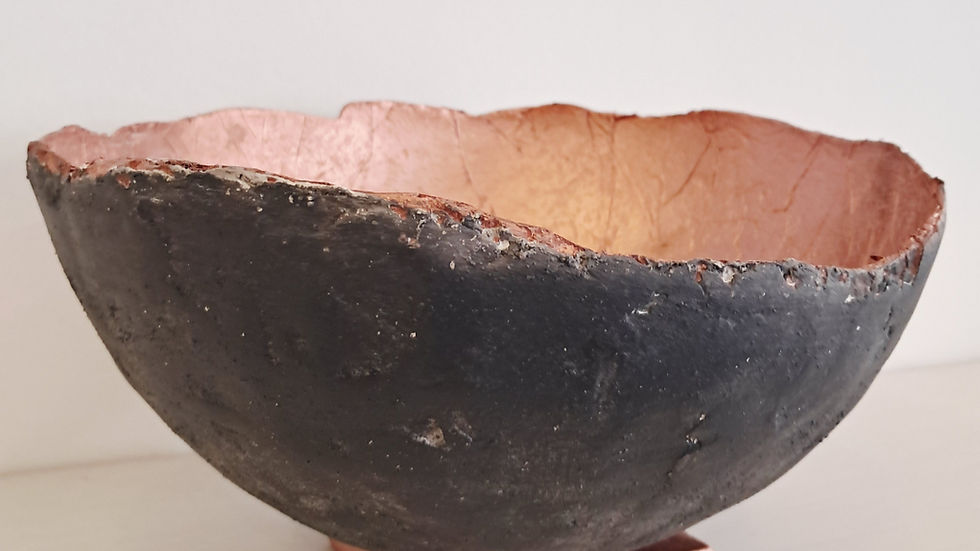 A textured black and copper bowl with a rough edge rests on a ceramic square base against a plain white background.