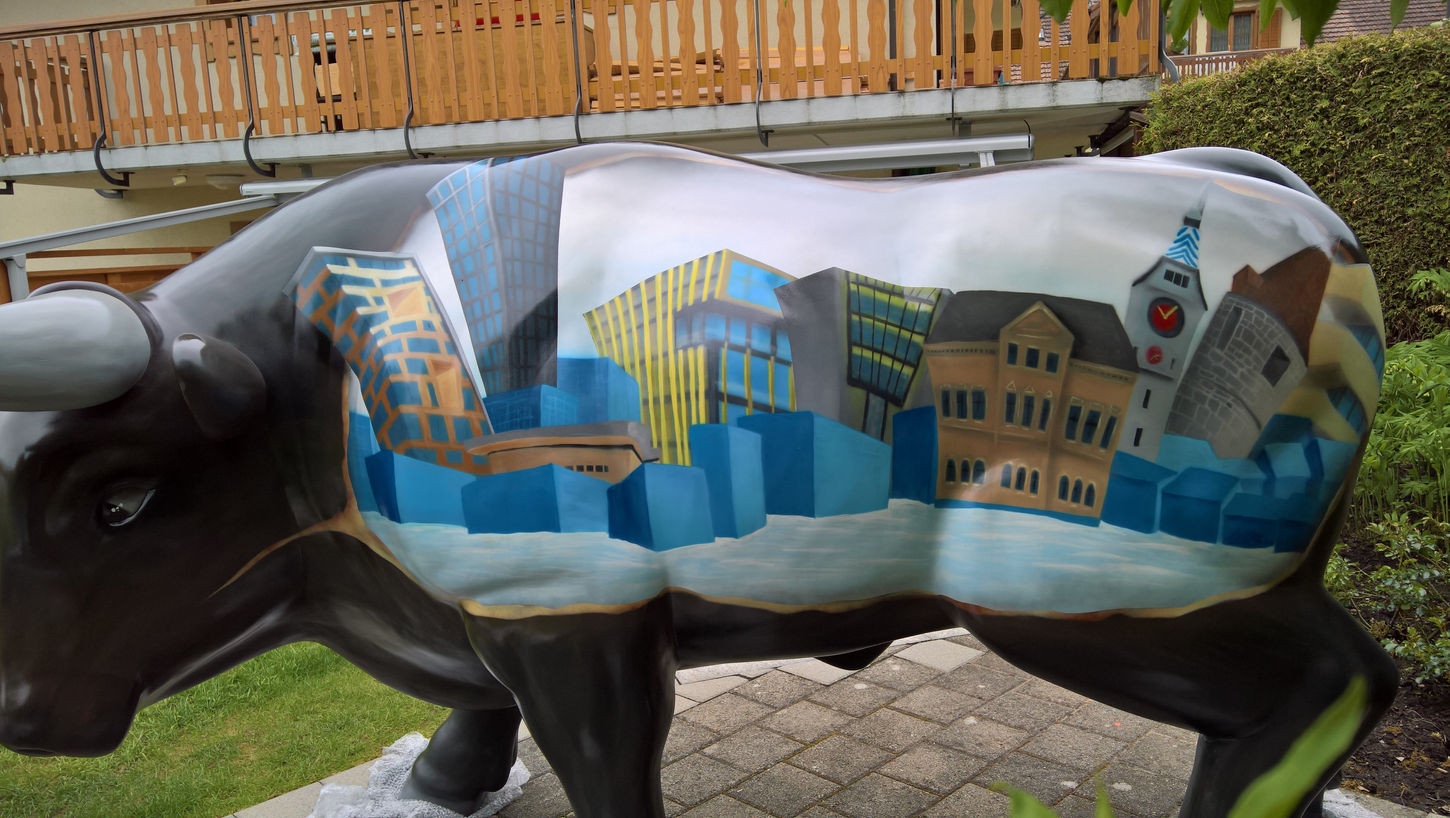 Die zweite Seite einer bemalten Stier-Skulptur, die eine Darstellung der Stadt Zug mit verschiedenen Gebäuden zeigt. Diese Skulptur wurde von dem Graffiti-Künstler SMOG für das Projekt 'Stierparade' zum 50. Jubiläum des EV Zug gestaltet.