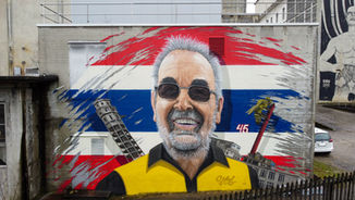 Mural zur Ehrung eines Mitarbeiters zu dessen Pensionierung und 45. Arbeitsjubiläum. Das Graffiti zeigt den Mitarbeiter als lächelnden Mann mit Sonnenbrille und grauem Bart vor einem Hintergrund in den Farben Rot, Weiß und Blau. Links im Bild ist ein schief stehender Turm zu sehen, rechts ein schräg stehender Schornstein mit einer Palme.