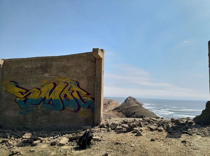 Ein Graffiti-Kunstwerk an einer alten Betonwand in Puerto Chicama, Peru, das den Schriftzug 'SMOG' in dynamischen, verschlungenen Buchstaben und leuchtenden Farben zeigt. Im Hintergrund ist ein Teil von 'la ola más larga del mundo' (die längste Welle der Welt) zu sehen. Das Bild wurde von dem Graffiti-Künstler SMOG während seiner Reise Südamerika Reise 2018 gestaltet.
