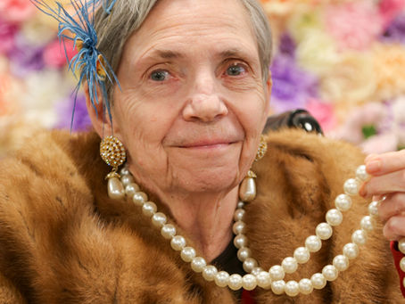 An older woman wearing large earrings, pearls, and fur shawl, smiling