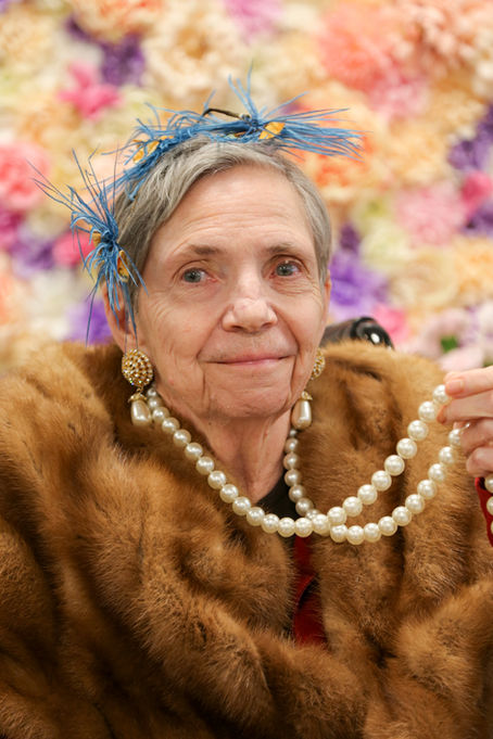 An older woman wearing large earrings, pearls, and fur shawl, smiling